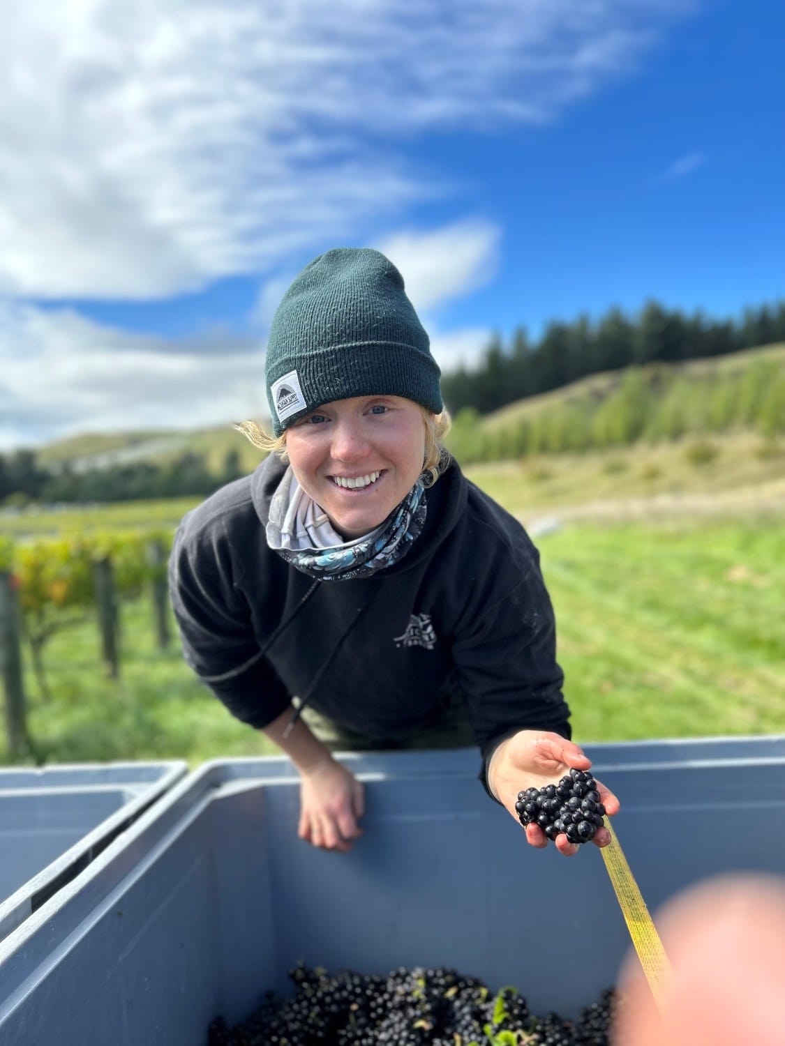 Lizzy wearing a green beanie, standing over grapes, holding grapes in hand.